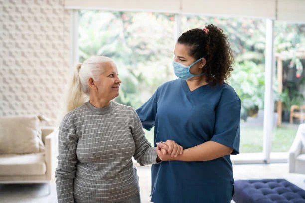 Home caregiver helping senior woman walking at home - wearing protecive face mask