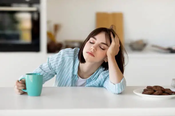 Sleepy young woman drinking coffee, feeling tired, suffering from insomnia and sleeping disorder. Sad female sitting in modern kitchen interior, empty space